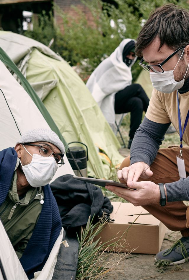 Outreach worker with badge crouching at tent and making notes in tablet while talking to refugee