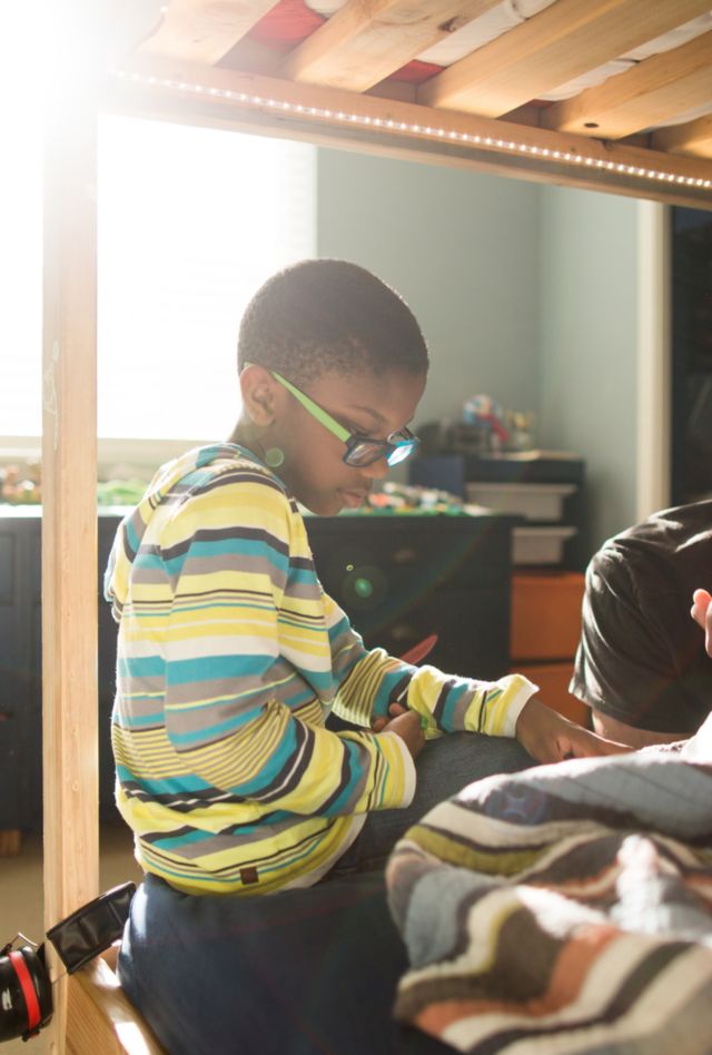 Black boy in glasses sits on a bunkbed in a child's room, reading a book. His father sits nearby, supervising.