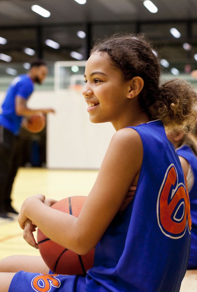 female young person holding a basketball  and sitting on the bench in an indoor basketball court with her teammates while the coach addresses the team