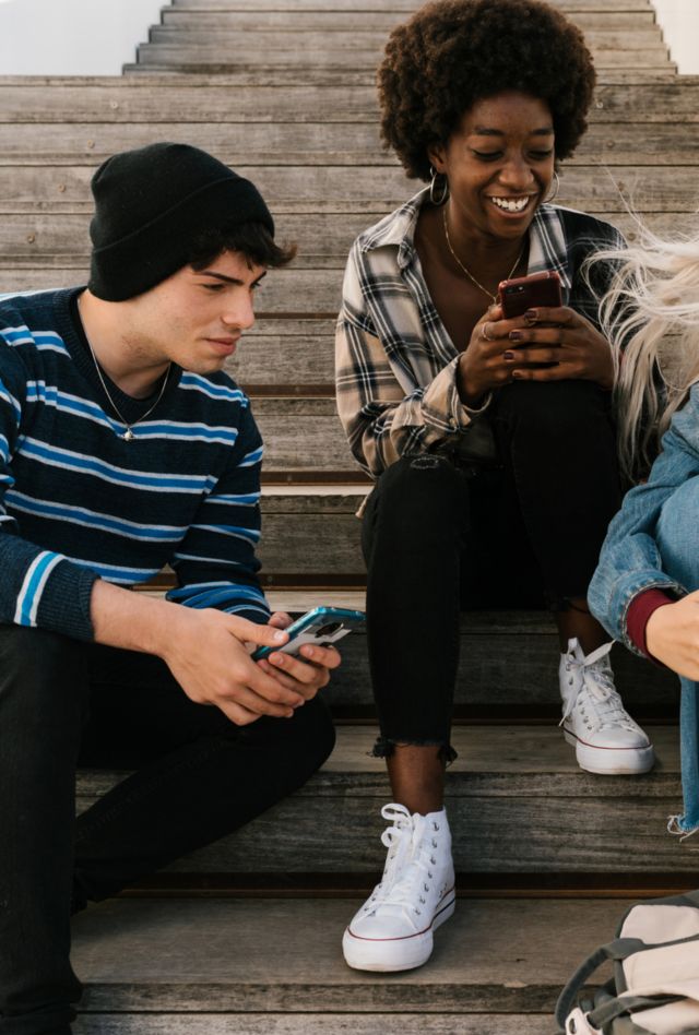 Happy diverse man and women smiling and browsing cellphones while resting on stairs on street