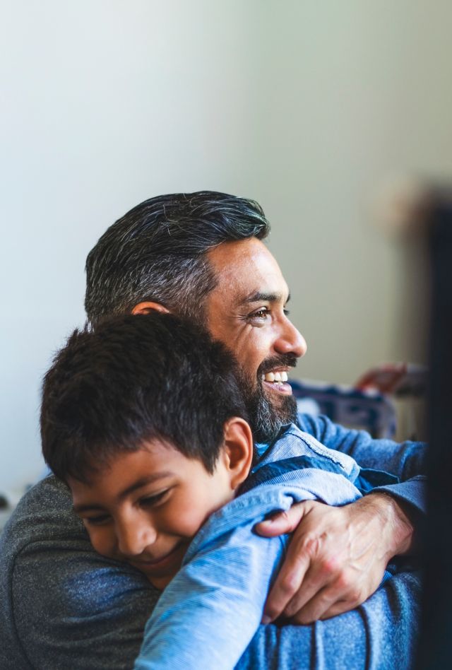 Smiling man embracing boy in bedroom. Happy father is looking away with son at home. They are spending leisure time.
