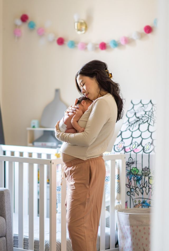 A new Mother stand in her daughters nursery as she takes a moment to enjoy Motherhood and soak it all in.  She is dressed comfortably and holding her baby close to her chest as the two bond.