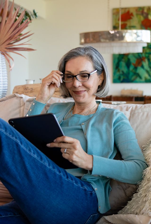 An older woman sits on her couch and uses her tablet.