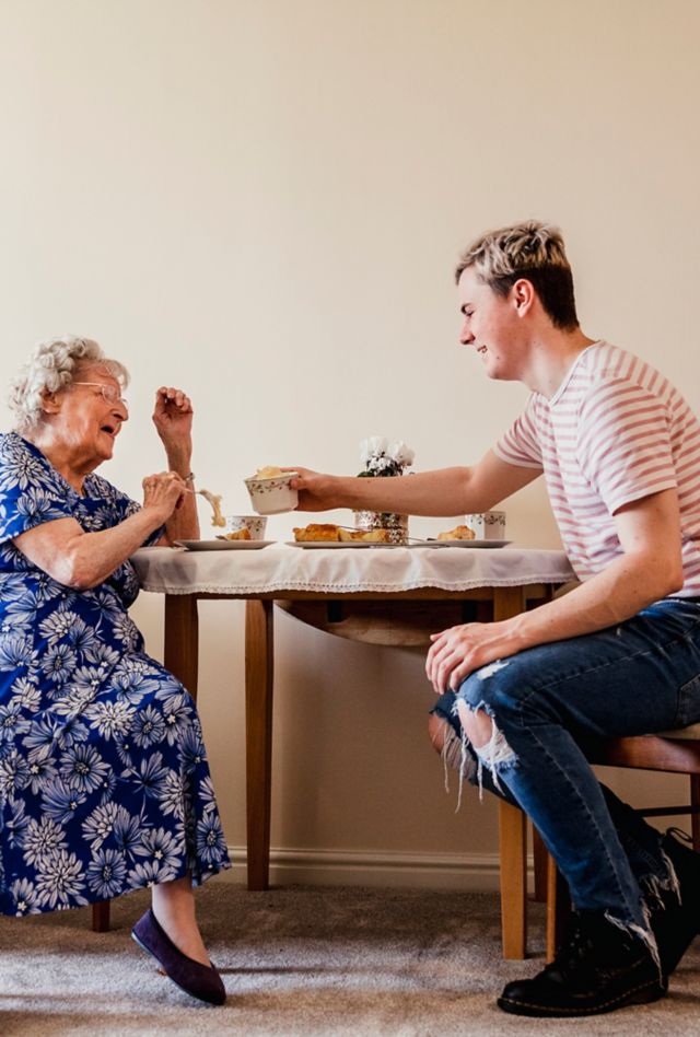 A young man sits having tea with an elderly woman. 