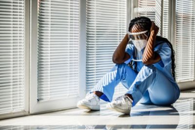 Wide shot of overworked, overwhelmed young nurse sitting on the floor, by the window, head in hands, eyes closed, having a short break from hard day at the hospital. She is wearing N95 face mask and face shield.