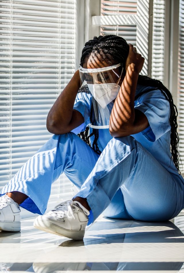 Wide shot of overworked, overwhelmed young nurse sitting on the floor, by the window, head in hands, eyes closed, having a short break from hard day at the hospital. She is wearing N95 face mask and face shield.