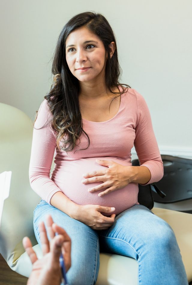 The mid adult pregnant woman touches her abdomen as she listens to the unrecognizable female doctor.  The doctor gestures as she holds the patient's medical records.