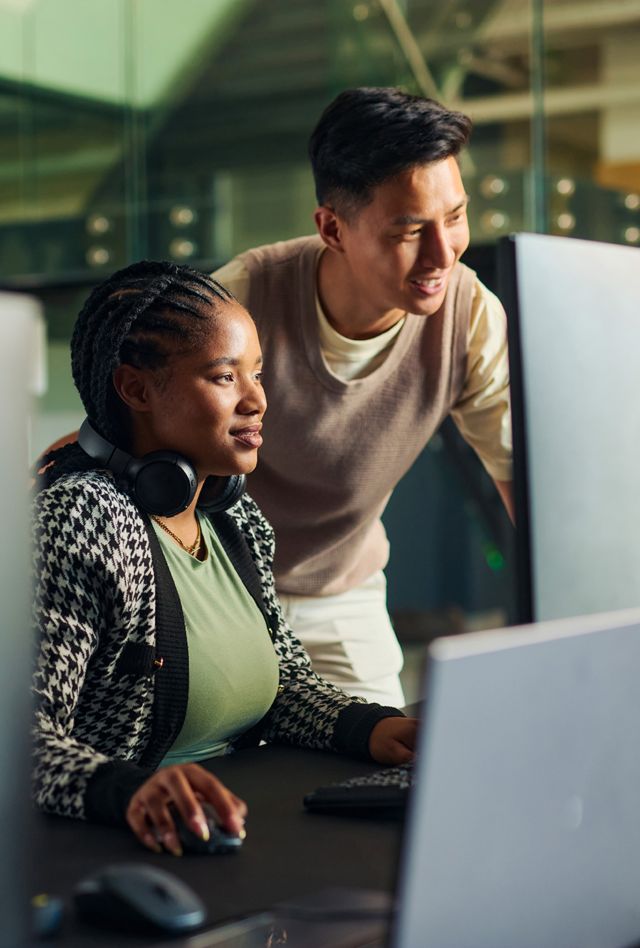 Two people work together at a computer workstation, with one seated at a desk wearing headphones and another standing beside them, looking at the monitor.