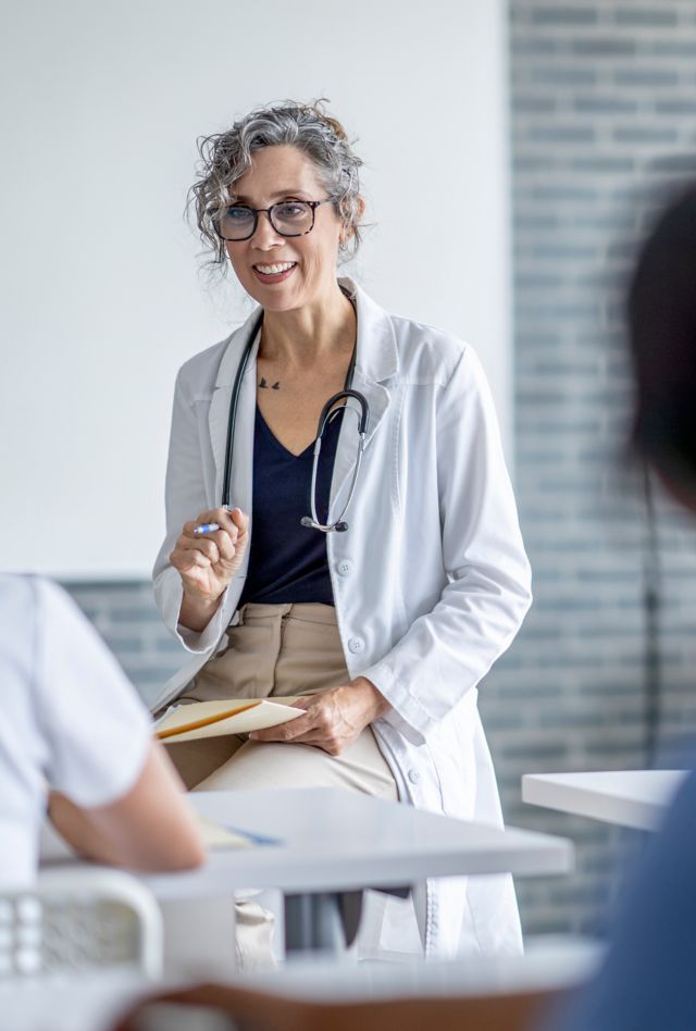 A mature female doctor sits at the front of a classroom as she teaches a small group of students.  She has a file folder on her lap as she talks with the students, and is dressed in a white lab coat.  The students are seated in front of her and paying close attention as they take notes.