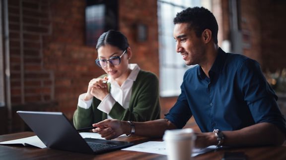 Two individuals seated at a table working together on a laptop, emphasizing teamwork and professionalism.