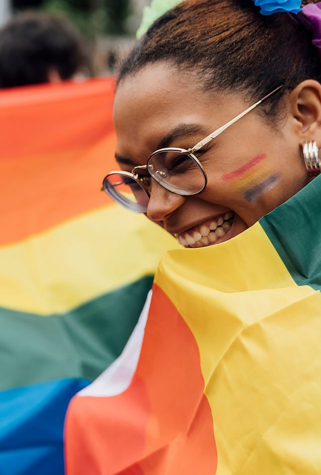 Young Beautiful Black Woman Covered with a Rainbow Pride Flag during the LGBTQ+ Festival in the City. The Street is crowded with Colourful people Enjoying for the event.