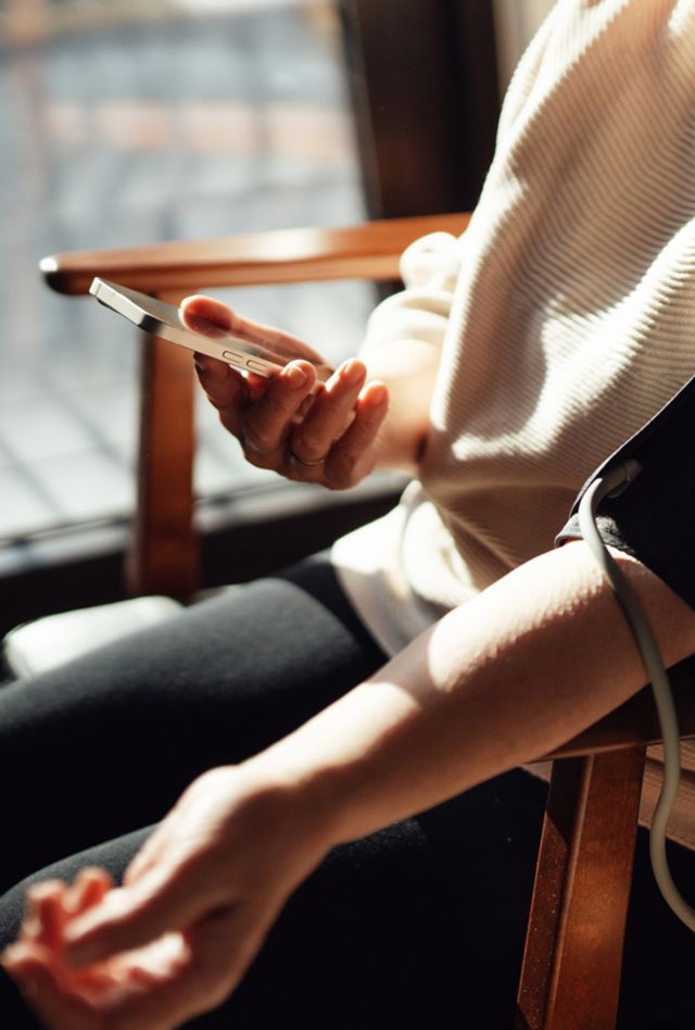 Person sitting in a chair wearing a blood pressure cuff and looking at their phone.