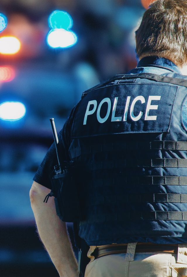 Blurry image of a police vehicle with lights flashing, and a person standing in foreground with back turned wearing a vest emblazoned with "Police" 