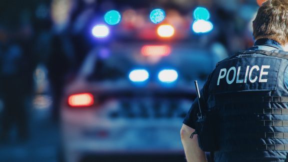 Foreground is the back of a person wearing a vest marked "POLICE," in background is blurred image of police car with flashing lights.