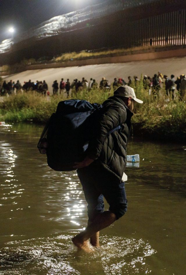 A man carrying a backpack walks across a river at night.  Other people are lined up along the other side of the river.