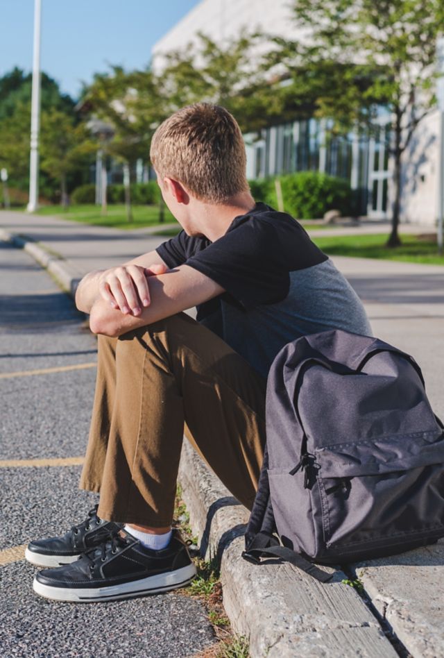 A teenage boy sitting on the curb in front of a school