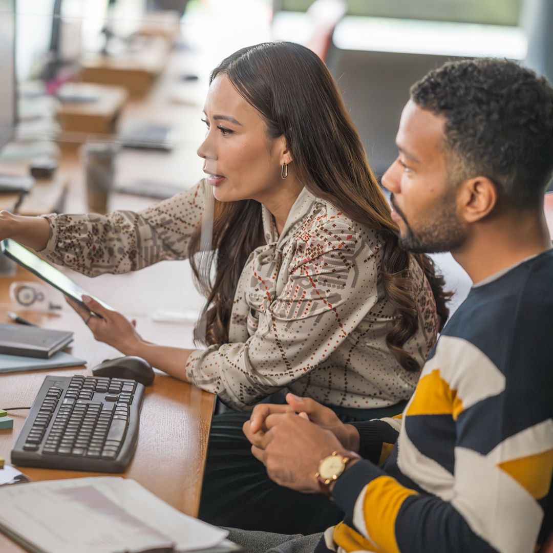 A woman and man analyze data on a desktop PC, engaged in conversation, wearing professional attire in a modern office setting.