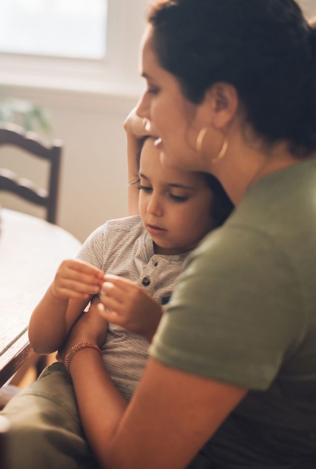 Sunlight hits the table as a woman cradles child in her lap while speaking with medical professional on a digital tablet. 