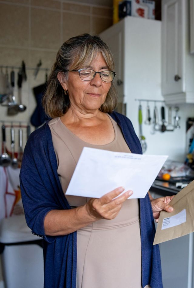 An older woman stands in a kitchen looking at mail