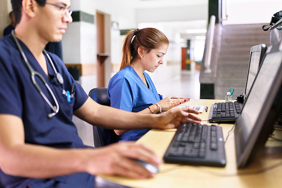 Two nurses in a hospital working at computers. The nurse in the foreground is wearing dark blue scrubs and the nurse in the background is wearing light blue scrubs.