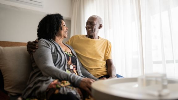 Older, dark-skinned couple smiling at each other on a couch in front of a sunlit curtain.