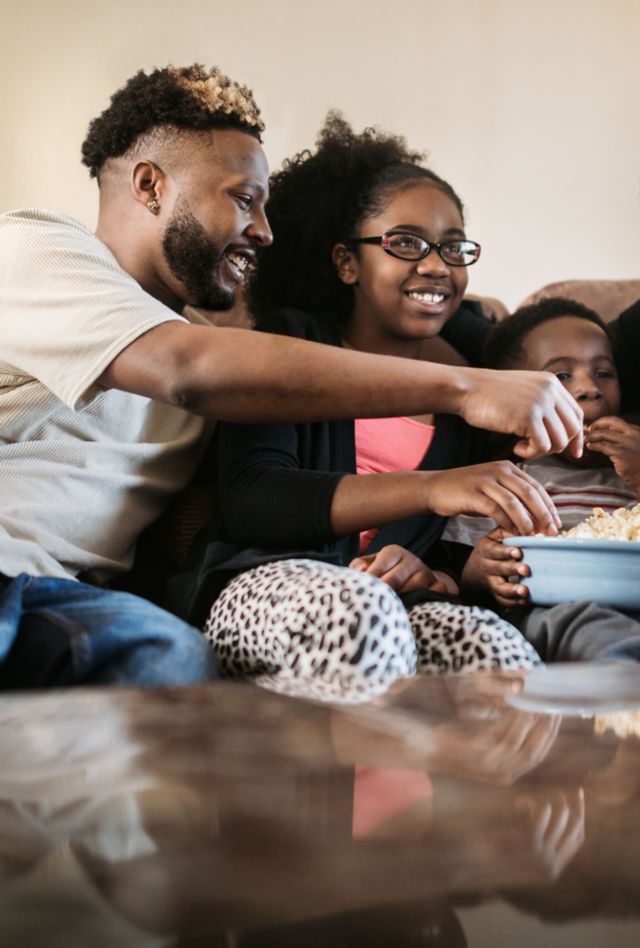 A fun African American family gets cozy on the sofa with a bowl of popcorn for their weekly movie tradition.  A time of connection and tenderness.