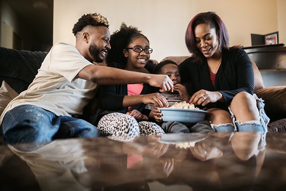 A fun African American family gets cozy on the sofa with a bowl of popcorn for their weekly movie tradition.  A time of connection and tenderness.