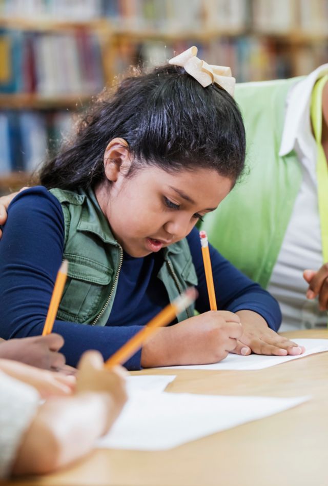 Elementary school students writing on paper with pencils at a table as their teacher looks on