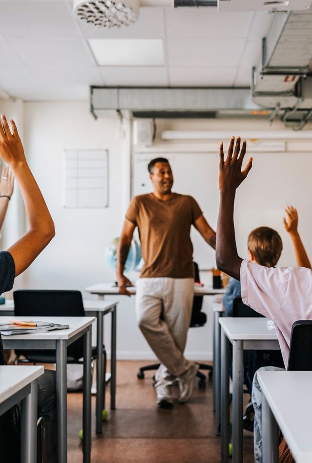 Teacher standing at the front of the class looking at students sitting in desks with their hands raised