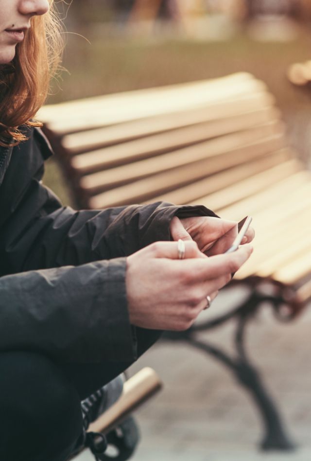 Young female in jacket and scarf sitting on wooden bench outdoors using smartphone