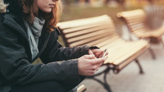 Young female in jacket and scarf sitting on wooden bench outdoors using smartphone