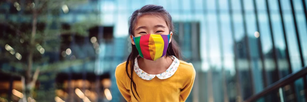 Une jeune fille portant un masque médical se penche vers la caméra et sourit joyeusement devant un immeuble de bureaux