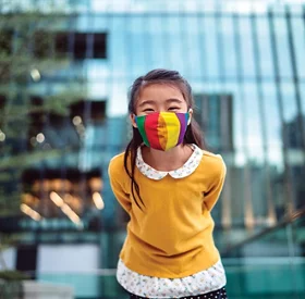 Une jeune fille portant un masque médical se penche vers la caméra et sourit joyeusement devant un immeuble de bureaux