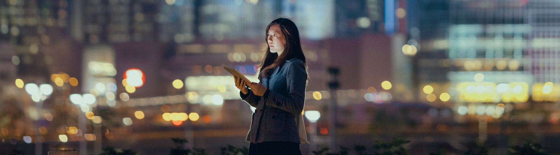Une jeune femme d’affaires utilise une tablette numérique dans un quartier financier, devant des gratte- ciel illuminés dans la nuit.