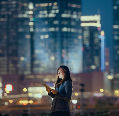 Une jeune femme d’affaires utilise une tablette numérique dans un quartier financier, devant des gratte- ciel illuminés dans la nuit.