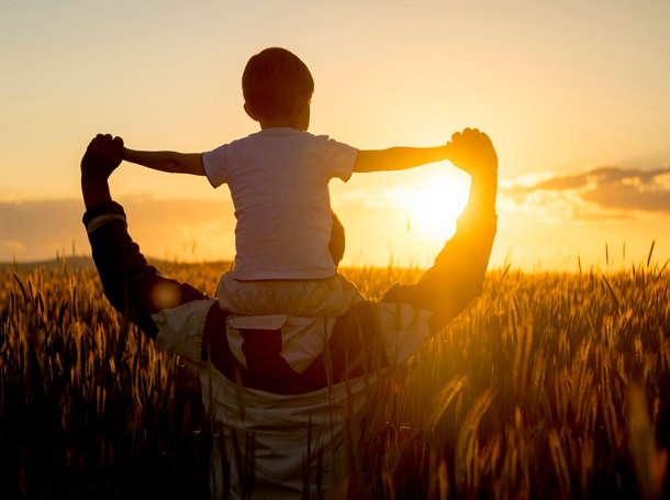 Father carrying son on shoulders in field of wheat at sunset