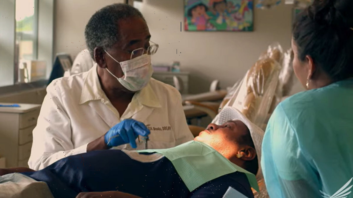 A doctor wearing a face mask speaks with a patient laying on an exam table,