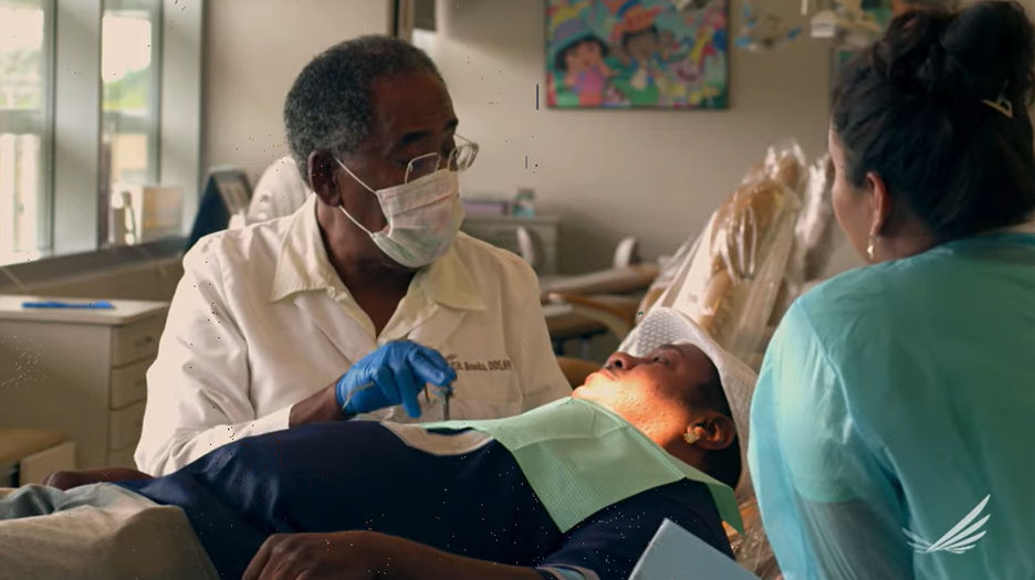 A doctor wearing a face mask speaks with a patient laying on an exam table,