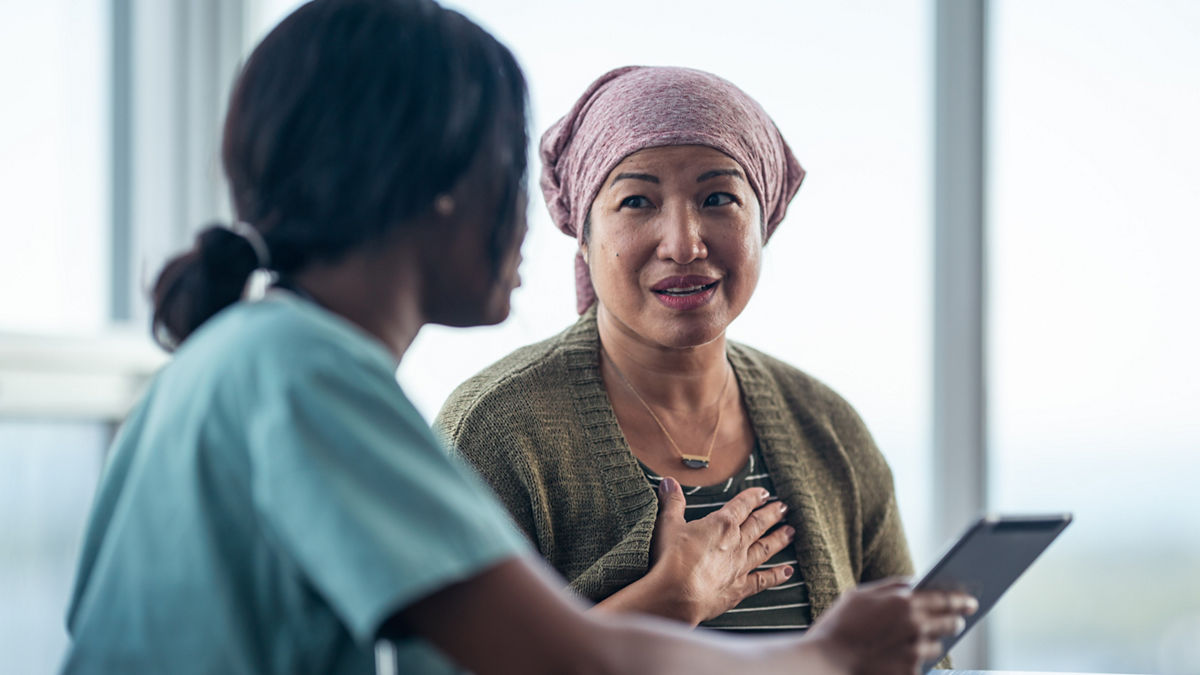 An Asian woman with cancer is consulting her doctor. The two women are seated at a table together. The patient is wearing a bandana to hide her hair loss. The medical professional is showing the patient test results on a digital tablet. They are discussing a treatment plan.