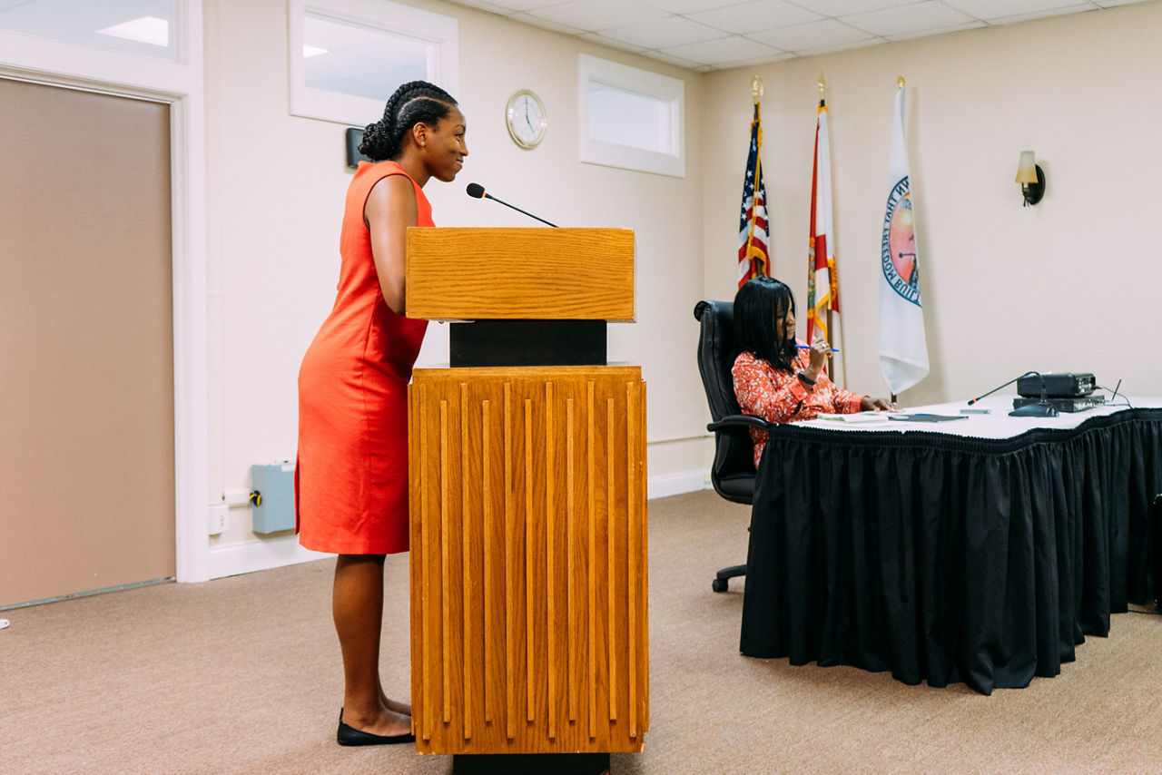 Side view of black woman age 20-28 speaking in microphone in a town hall meeting with Asian woman sitting at table next to her. 