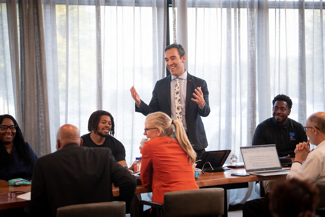 A man stands while speaking to a room full of people gathered around a table. 