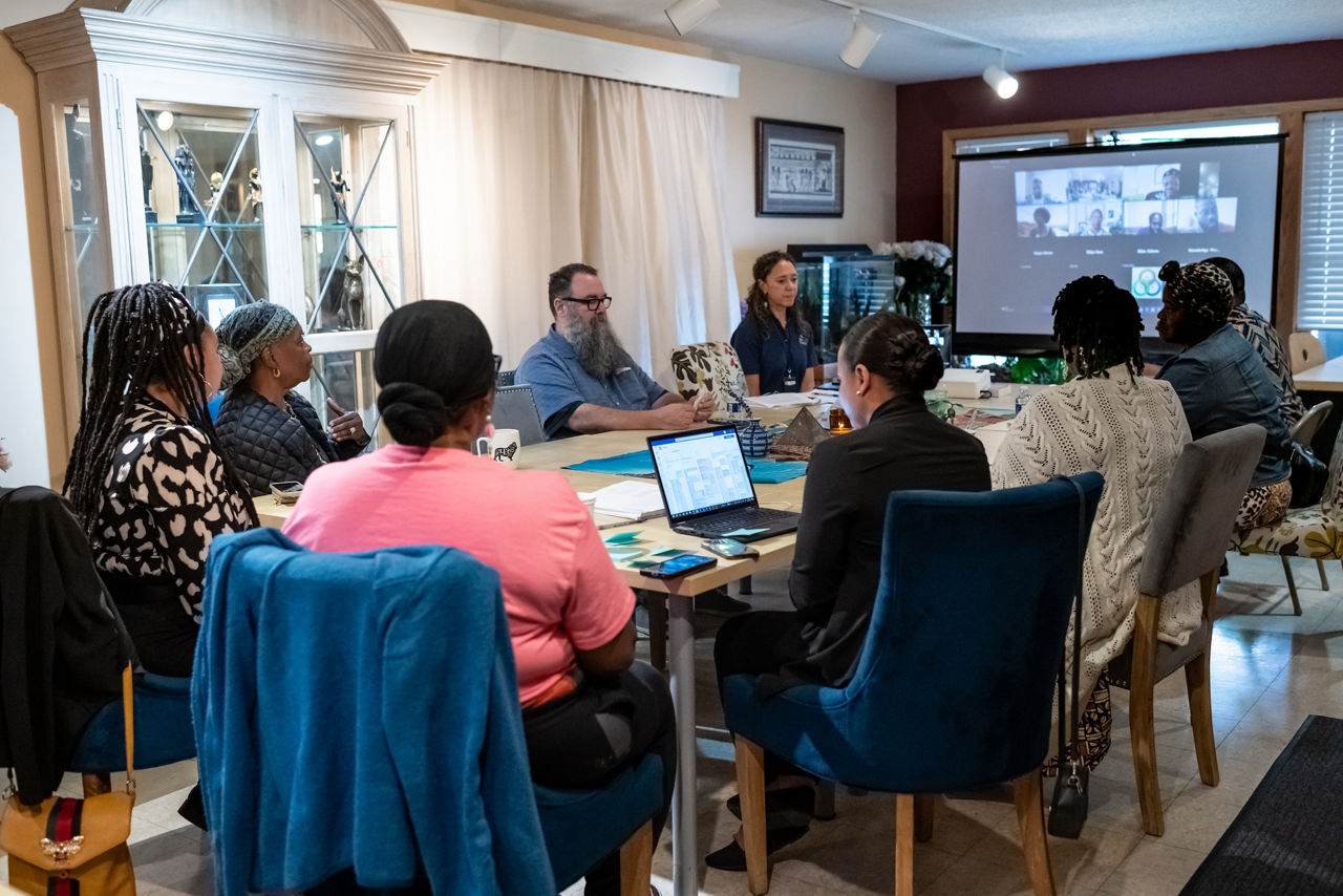 : a group of people gathering around a conference table. A projector screen is on the wall showing virtual attendees via Zoom.