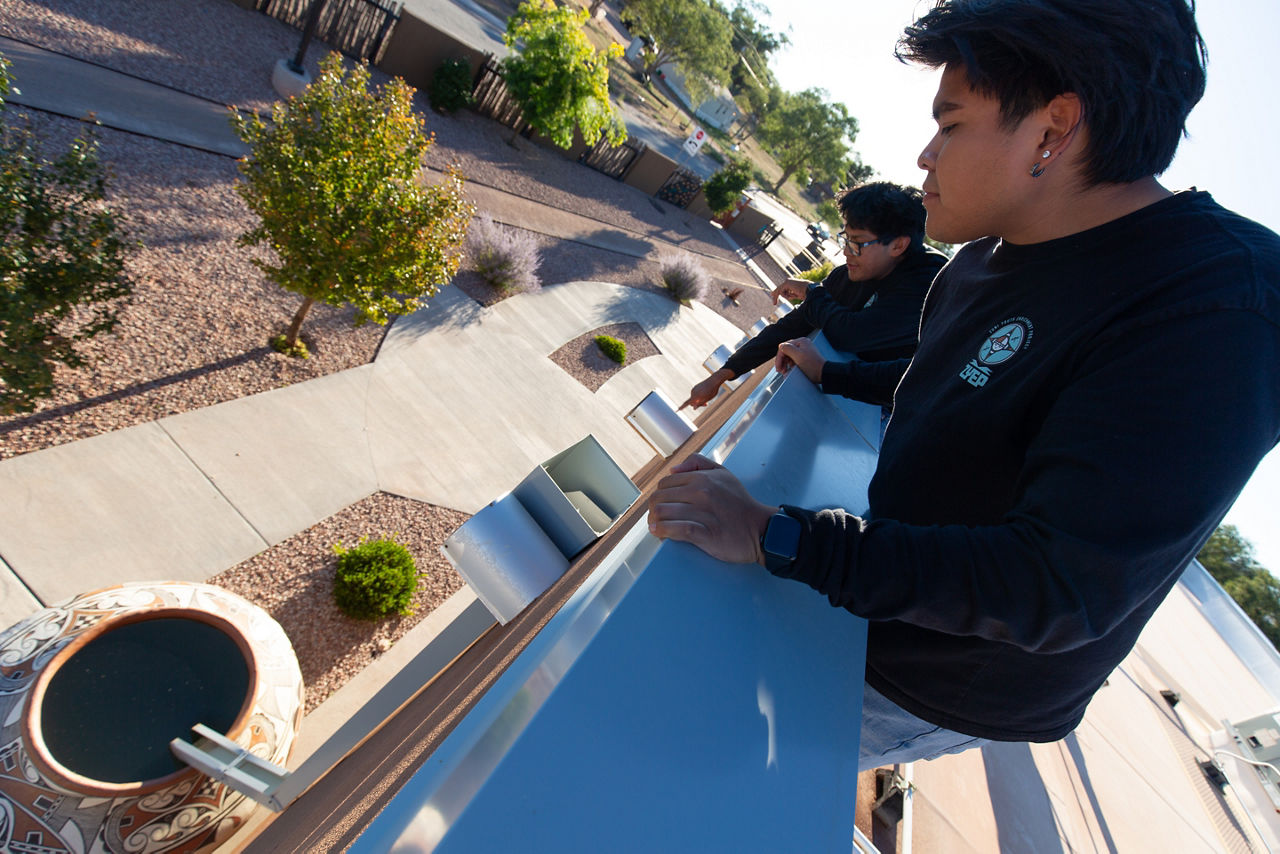 Zuni Youth Enrichment Project (ZYEP) Food Sovereignty team members, Zachary James (back) shows Dylan Solomon, Jr (front) the rainwater collection system at the ZYEP building, which collects water off the roof and stores it in large clay pots that are decorated by local artists, in order to water the gardens on the property. Water catchment systems are essential for food cultivation/plant growth in the Zuni climate.