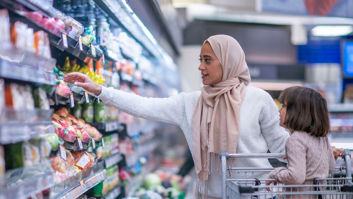 A young Muslim mother and daughter walk the produce aisles of the grocery store together shopping for groceries.  They are both dressed casually and the mother is wearing a Hijab.  The mother is reaching for products on the shelf while pushing a shopping cart with her daughter seated in it.