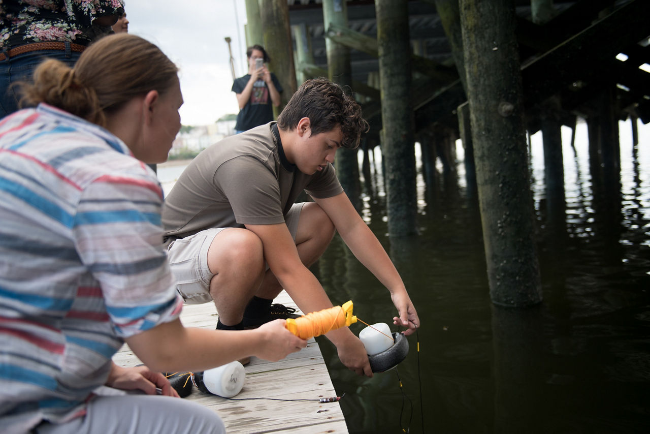 Christian Garcia, center, 17, a youth member of GreenRoots, and researcher Sara Wylie, left, with Public Lab, prepare a water testing kit for placement in the Chelsea Creek. The kit will be used to assess temperature impacts of industrial waste product discharge into the creek. (AEM Chelsea story image)