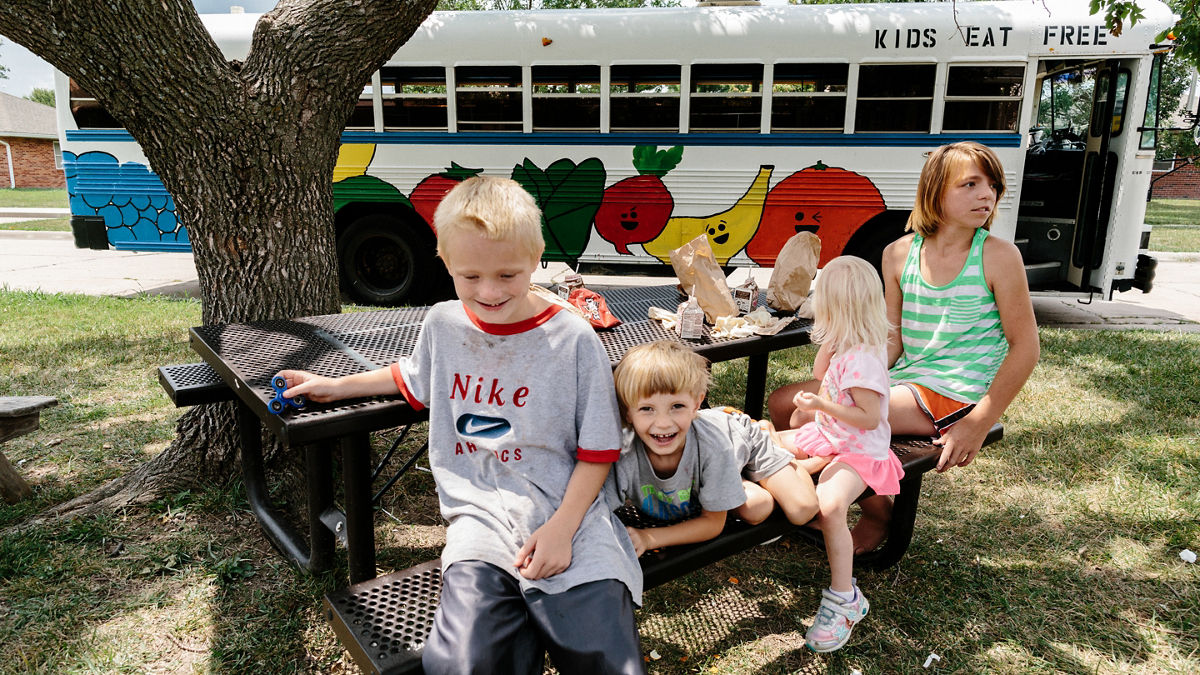 Iola, KS - August 1, 2017 - Local youth (L to R) Conner McCoullough, Ryan Cress, Lilly Cunningham, and Katlin Cress eat lunch at a picnic table in front of the MARV ("Meals And Reading Vehicle") bus in a residential neighborhood in Iola.