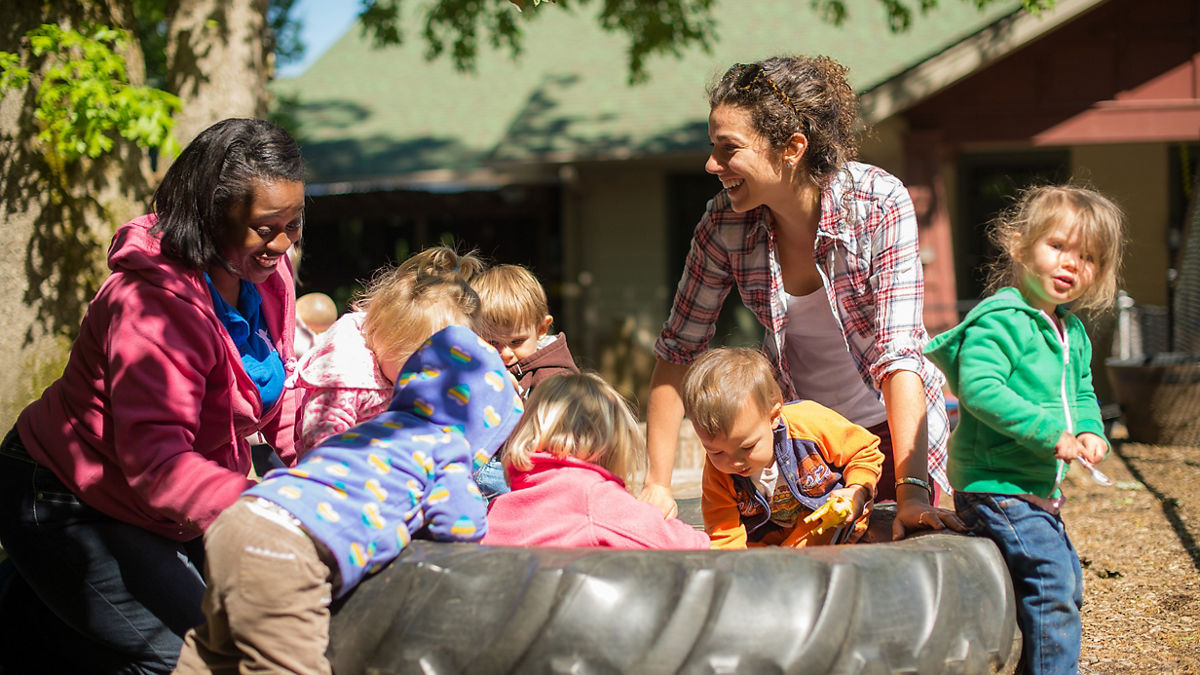 A daycare teacher playing with a group of young children outside.