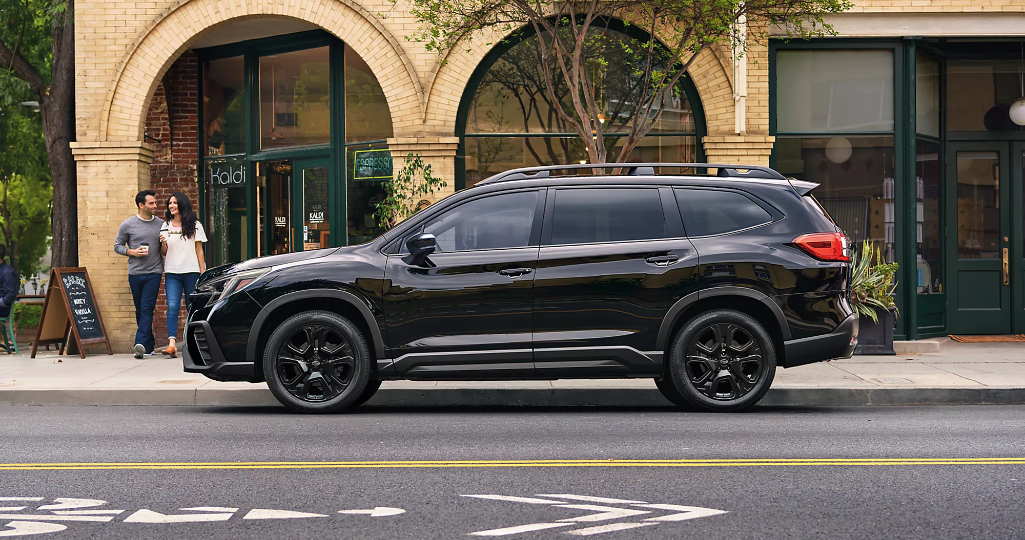 Black 2024 Subaru Ascent with black metal alloy wheels parallel parked in front of a coffee shop with a family walking out
