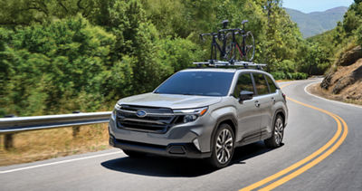 A 2025 Forester with two bicycles on its roof rack drives through trees and rolling hills.