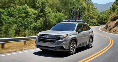 A 2025 Forester with two bicycles on its roof rack drives through trees and rolling hills.